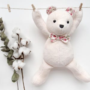 May include: A pink plush teddy bear with a floral bow tie hanging on a clothesline with cotton and eucalyptus branches on either side.