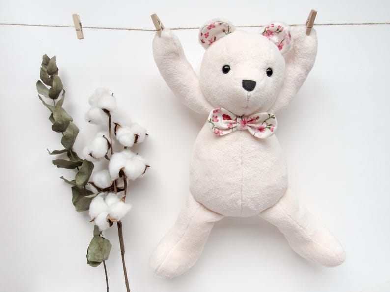 May include: A pink plush teddy bear with a floral bow tie hanging on a clothesline with cotton and eucalyptus branches on either side.