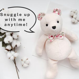 K&ouml;nnte beinhalten: Ein weicher, wei&szlig;er Pl&uuml;schteddyb&auml;r mit einer floralen Schleife. Der B&auml;r sitzt auf einer wei&szlig;en Oberfl&auml;che mit einer Sprechblase, auf der "Snuggle up with me anytime!" steht.