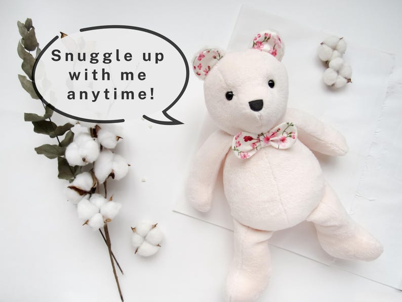 K&ouml;nnte beinhalten: Ein weicher, wei&szlig;er Pl&uuml;schteddyb&auml;r mit einer floralen Schleife. Der B&auml;r sitzt auf einer wei&szlig;en Oberfl&auml;che mit einer Sprechblase, auf der "Snuggle up with me anytime!" steht.