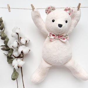 A cute handmade plush teddy bear hanging from a clothesline, serving as a perfect example of a memory bear pattern or teddy bear pattern. This bear sewing pattern features soft fabric with floral accents on the ears and bow tie, making it a wonderful DIY memorial gift or DIY memorial bear. This design also works well as a bunny sewing pattern or general plush sewing pattern.