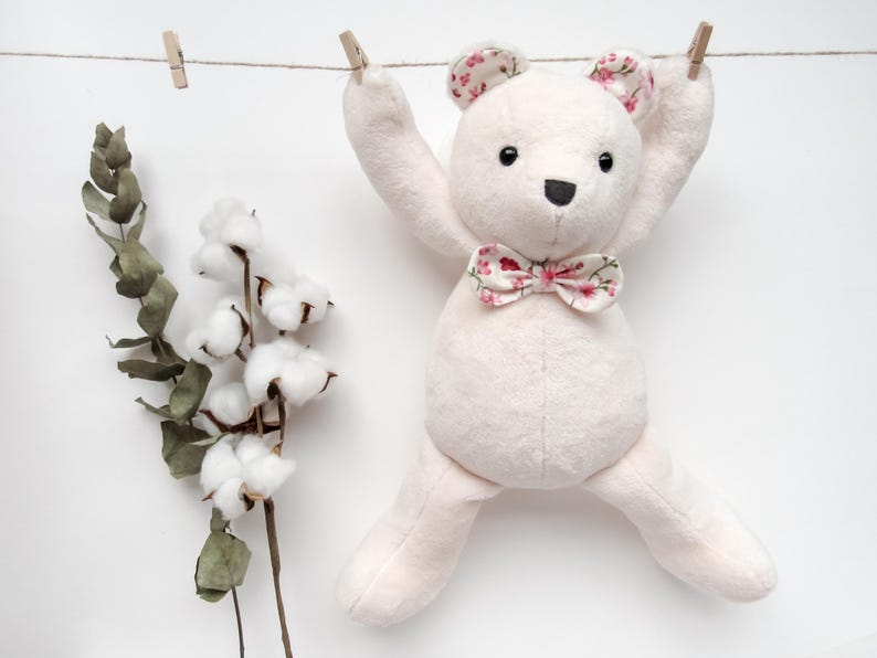 A cute handmade plush teddy bear hanging from a clothesline, serving as a perfect example of a memory bear pattern or teddy bear pattern. This bear sewing pattern features soft fabric with floral accents on the ears and bow tie, making it a wonderful DIY memorial gift or DIY memorial bear. This design also works well as a bunny sewing pattern or general plush sewing pattern.