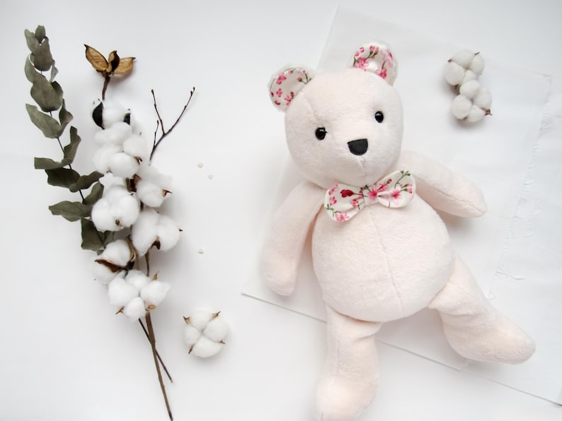 May include: A plush pink teddy bear with floral print ears and a bow tie. The bear is sitting on a white surface with cotton balls and a branch with cotton balls.
