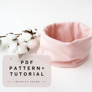 May include: A soft pink infinity scarf, displayed on a white surface, with a sprig of cotton blossoms. The scarf is a tube shape, and the image includes text that reads "PDF PATTERN + TUTORIAL - INFINITY SCARF."