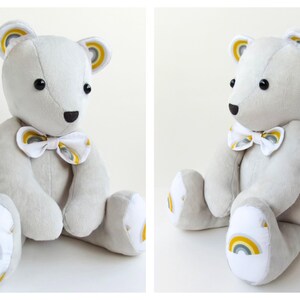 K&ouml;nnte beinhalten: Ein hellgrauer Pl&uuml;schteddyb&auml;r mit schwarzen Knopfaugen und einer schwarzen Nase. Der B&auml;r tr&auml;gt eine wei&szlig;e Fliege mit einem gelb-grauen Regenbogenmuster. Passende Regenbogenmotive befinden sich an den Innenohren und den Fu&szlig;sohlen.