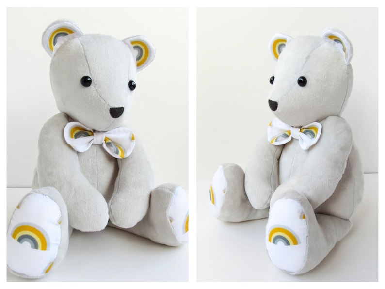 K&ouml;nnte beinhalten: Ein hellgrauer Pl&uuml;schteddyb&auml;r mit schwarzen Knopfaugen und einer schwarzen Nase. Der B&auml;r tr&auml;gt eine wei&szlig;e Fliege mit einem gelb-grauen Regenbogenmuster. Passende Regenbogenmotive befinden sich an den Innenohren und den Fu&szlig;sohlen.