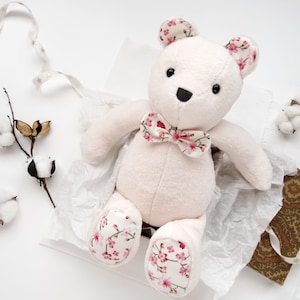 May include: A soft, white plush teddy bear with a pink floral bow tie and matching floral print on the paws. The bear is sitting on a white tissue paper background.