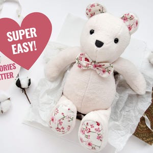 K&ouml;nnte beinhalten: Ein wei&szlig;er Pl&uuml;schteddyb&auml;r mit einer rosa floralen Schleife und rosa floralen Akzenten an den Pfoten. Der B&auml;r sitzt auf einer wei&szlig;en Oberfl&auml;che mit einem herzf&ouml;rmigen Schild, auf dem "Super Easy!" steht, und einem Schild, auf dem "Memories Matter" steht.