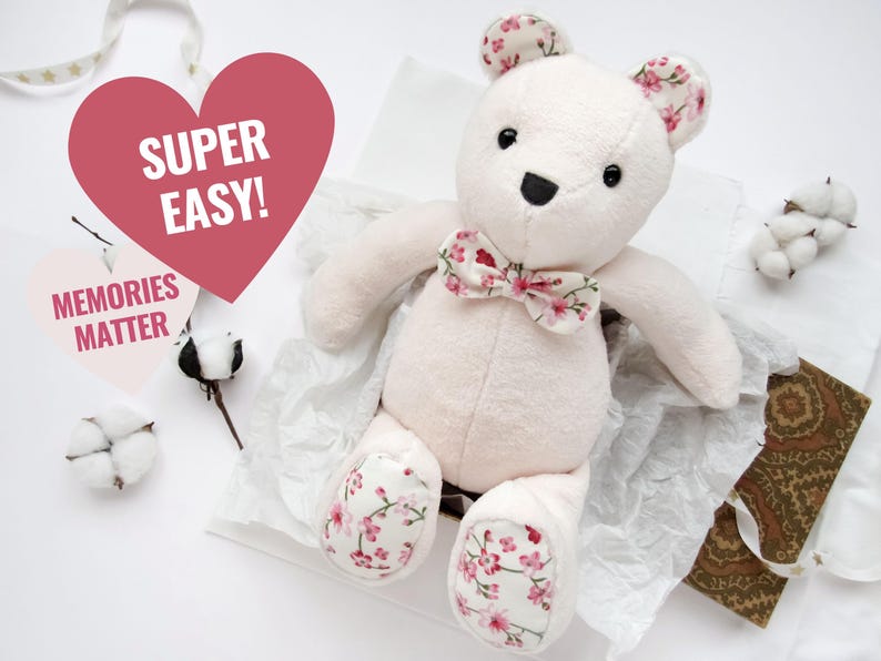 K&ouml;nnte beinhalten: Ein wei&szlig;er Pl&uuml;schteddyb&auml;r mit einer rosa floralen Schleife und rosa floralen Akzenten an den Pfoten. Der B&auml;r sitzt auf einer wei&szlig;en Oberfl&auml;che mit einem herzf&ouml;rmigen Schild, auf dem "Super Easy!" steht, und einem Schild, auf dem "Memories Matter" steht.