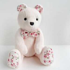 A cute handmade plush teddy bear made from soft light pink fabric with floral fabric accents on its ears, bowtie, and foot pads. This memory bear pattern is perfect as a DIY memorial gift. This easy sewing pattern for a stuffed bear pattern is available as plushie patterns PDF. Whether you are looking for a teddy bear sewing pattern or bunny sewing pattern, this DIY is a wonderful cute animal pattern that fits perfectly among toy sewing patterns and memory bear template projects.