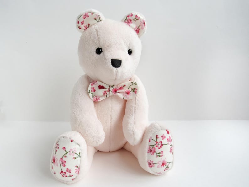 A cute handmade plush teddy bear made from soft light pink fabric with floral fabric accents on its ears, bowtie, and foot pads. This memory bear pattern is perfect as a DIY memorial gift. This easy sewing pattern for a stuffed bear pattern is available as plushie patterns PDF. Whether you are looking for a teddy bear sewing pattern or bunny sewing pattern, this DIY is a wonderful cute animal pattern that fits perfectly among toy sewing patterns and memory bear template projects.