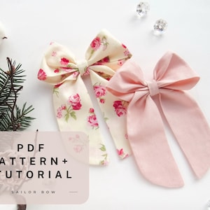 Peut inclure: Deux nœuds en tissu pour cheveux, l'un à imprimé floral et l'autre en rose uni. Les nœuds sont noués avec de longues queues et sont présentés sur une surface blanche. Le texte "PDF PATTERN + TUTORIAL SAILOR BOW" est visible sur l'image.
