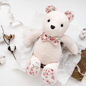 A handmade plush teddy bear made from a memory bear pattern, sitting in a gift box lined with tissue paper. This cute animal pattern features soft, light-colored fabric with floral-patterned accents on the ears, bowtie, and paw pads. Perfect as a DIY memorial gift, this easy sewing pattern is ideal for creating a custom DIY memorial bear or a thoughtful keepsake. This project is a great example of versatile toy sewing patterns, including styles similar to a bunny sewing pattern.