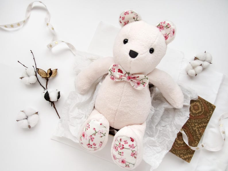 A handmade plush teddy bear made from a memory bear pattern, sitting in a gift box lined with tissue paper. This cute animal pattern features soft, light-colored fabric with floral-patterned accents on the ears, bowtie, and paw pads. Perfect as a DIY memorial gift, this easy sewing pattern is ideal for creating a custom DIY memorial bear or a thoughtful keepsake. This project is a great example of versatile toy sewing patterns, including styles similar to a bunny sewing pattern.