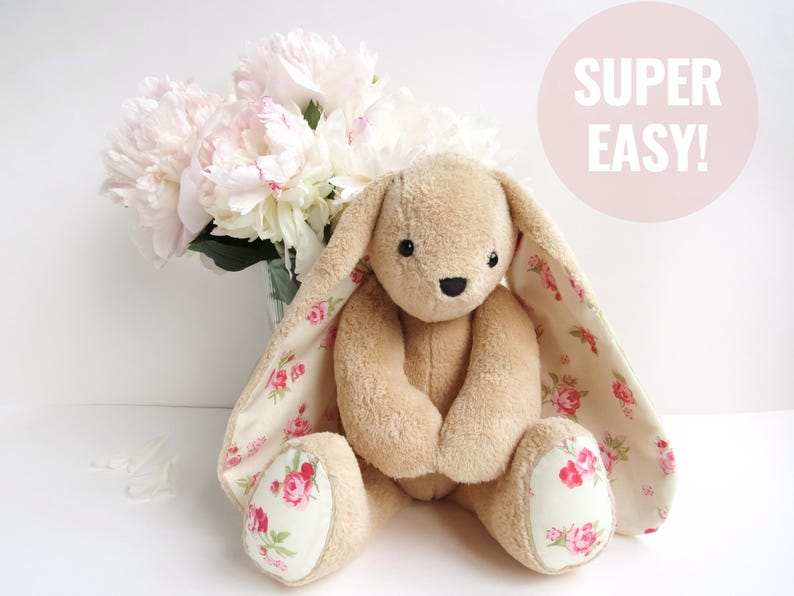 A cute, handmade stuffed bunny plushie sitting in front of a bouquet of peonies. The toy is made from light brown plush fabric with floral fabric accents on its long ears and feet. It is a versatile plush sewing pattern that serves as a bunny sewing pattern, a stuffed bear pattern, or a bear sewing pattern. Perfect for those looking for plushie patterns pdf or toy sewing patterns to make a DIY memorial bear or simply a charming cute animal pattern.
