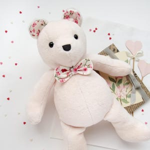 May include: A pink plush teddy bear with a floral bow tie. The bear is sitting on a white surface with pink hearts scattered around it.