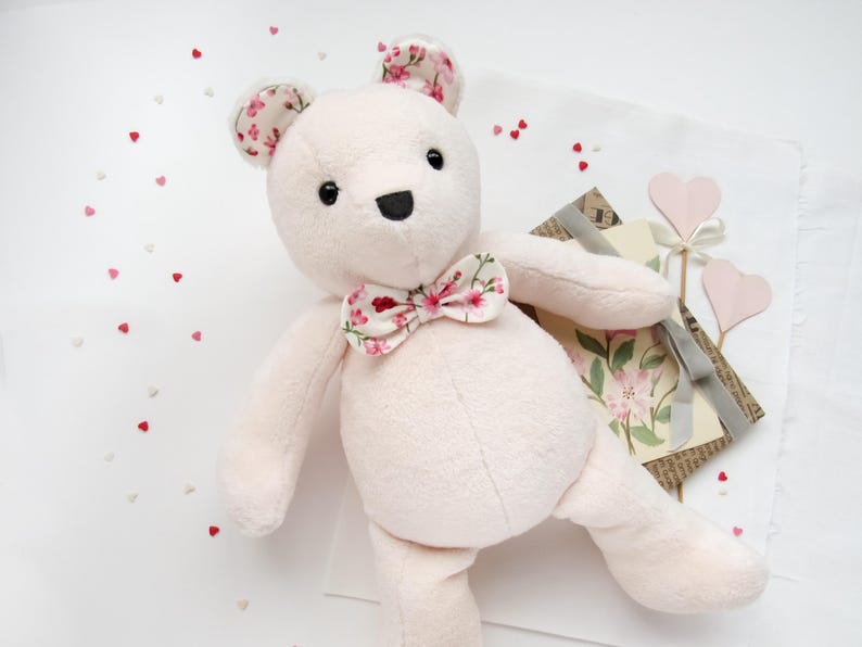 May include: A pink plush teddy bear with a floral bow tie. The bear is sitting on a white surface with pink hearts scattered around it.