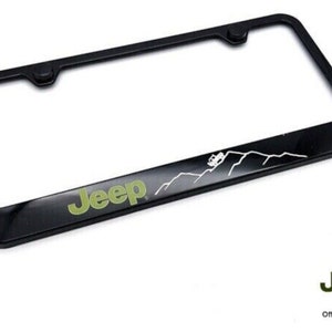 May include: Black metal license plate frame with a green Jeep logo and a white silhouette of a Jeep driving over mountains.