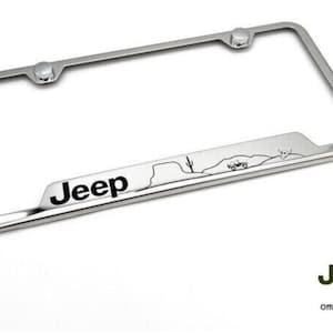 May include: Chrome license plate frame with a black Jeep logo and a silhouette of a Jeep driving through a desert landscape. The frame has two silver mounting posts.