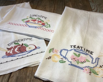 May include: Three white kitchen towels with embroidered designs and text. The top towel says "Lunchtime" and features a bowl of fruit. The middle towel says "Dinnertime" and features an apple on a plate. The bottom towel says "Teatime" and features a teapot with flowers.