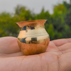 Puede incluir: Una pequeña olla de latón antigua con un borde acampanado y un cuerpo redondeado. La olla se sostiene en una mano, con un fondo verde borroso. La superficie de la olla refleja el entorno, creando un tono dorado cálido.