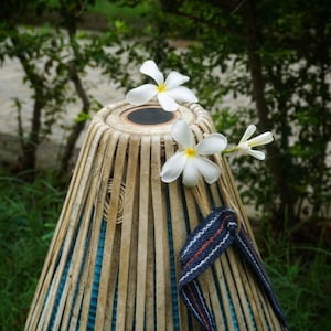 Puede incluir: Un tambor tabla indio tradicional con un acabado de madera natural, adornado con flores blancas de plumeria. El tambor tiene una correa azul y blanca, sobre un fondo de follaje verde.