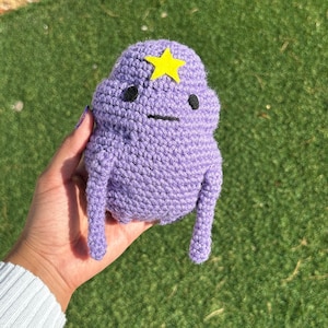 May include: A purple crocheted character with a yellow star on its head. The character has black eyes and a small mouth.