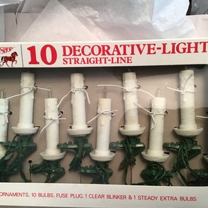 Puede incluir: Una caja de 10 luces navideñas blancas en forma de vela con cable verde. La caja dice "10 DECORATIVE-LIGHTS STRAIGHT-LINE" y "10 ORNAMENTS, 10 BULBS, FUSE PLUG, 1 CLEAR BLINKER & 1 STEADY EXTRA BULBS".