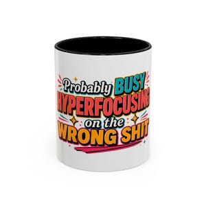 May include: A white ceramic mug with a black interior and rim. The exterior features colorful, bold text that reads: Probably BUSY HYPERFOCUSING on the WRONG SHIT, accented with small star and dash graphics in shades of red, blue, yellow, and teal.
