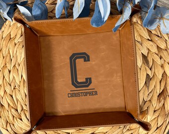 May include: A square, brown leather valet tray with a dark gray letter "C" and the name "Christopher" printed on the inside. The tray is on a woven, tan surface with blue decorative leaves.