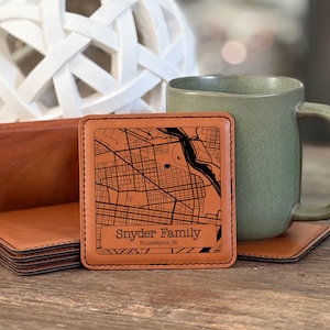 May include: A square, tan leather coaster with a black map design and the text "Snyder Family, Philadelphia, PA." A green mug and a stack of coasters are in the background. The coaster is approximately 4 inches square.