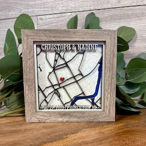 May include: A framed map art piece with a layered design. The map features a red heart marking a location, a blue river, and the names "CHRISTOPH & NADINE" above the map. The date "06.27.1999 PRINCETON, NJ" is printed below the map.
