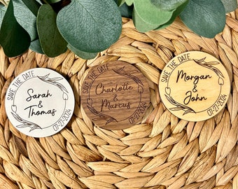 May include: Three round wooden save the date magnets with a floral design and the names of the couples and their wedding date. The magnets are on a woven wicker surface.