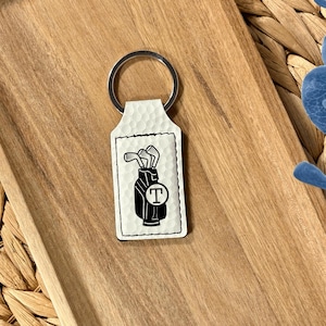 May include: A white rectangular keychain with a dimpled texture featuring a black graphic of a golf bag containing clubs and a circular emblem with the letter T in the center. The keychain includes a silver metal split ring at the top.