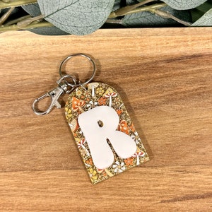 May include: A white letter R keychain with a gold glitter background and a mushroom pattern. The keychain has a silver metal ring and clasp.