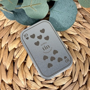 May include: A rectangular silver metal tin with rounded corners featuring a printed design of ten dark gray hearts, the number 10 in two corners, and the text Happy tin Anniversary centered on the lid in a dark gray sans-serif font.