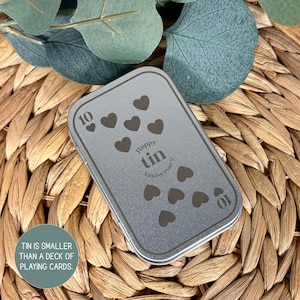 May include: A rectangular silver-toned metal tin with rounded corners. The lid features a dark gray printed design of ten heart shapes, the number 10 in two corners, and the text Happy Tin Anniversary in the center. The tin is smaller than a deck of playing cards.