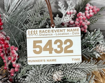 May include: A white wooden race bib ornament with brown text and a brown leather cord for hanging. The ornament features the number 5432, and text that reads "RACE/EVENT NAME, Distance - Location - Date, RUNNER'S NAME, TIME."