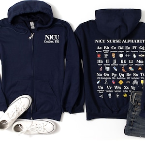May include: A navy blue zippered hoodie with a white graphic on the back that says "NICU Nurse Alphabet" and includes an alphabet with icons representing medical terms related to the NICU. The left side of the hoodie has a white graphic that says "NICU Custom. RN".