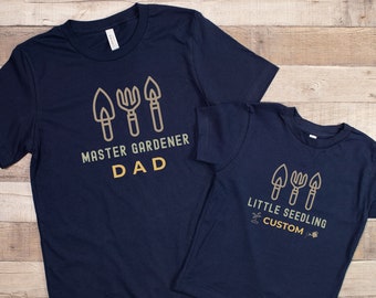 May include: Two navy blue t-shirts with a gardening theme. The larger shirt says "Master Gardener Dad" with a graphic of gardening tools. The smaller shirt says "Little Seedling Custom" with the same gardening tool graphic.