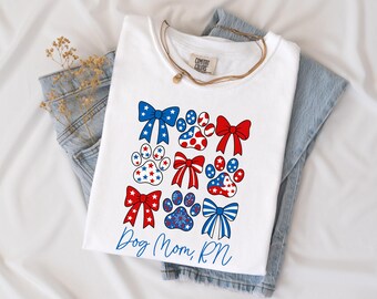May include: White t-shirt with a patriotic design featuring paw prints and bows in red, white, and blue. The shirt includes the text "Dog Mom, RN" in blue script. The shirt is folded on top of a pair of blue jeans.