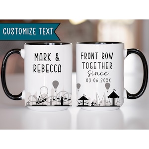May include: Two white coffee mugs with black handles and black rims. The mug on the left says "Mark & Rebecca". The mug on the right says "Front Row Together Since 03.06.20XX". Both mugs feature a black and white silhouette of a carnival or amusement park.