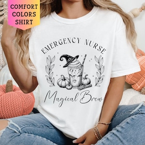 May include: White t-shirt with black text that reads "Emergency Nurse Magical Brew". The text is surrounded by a black and white illustration of a witch's hat, a pumpkin, and a cup of coffee.
