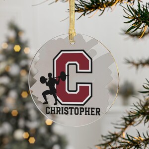May include: A clear glass Christmas ornament with a red and white letter C and a silhouette of a person lifting weights. The ornament has the name "CHRISTOPHER" printed below the letter.