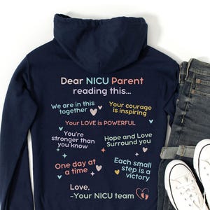 May include: Navy blue hoodie with the text "Dear NICU Parent reading this..." and supportive messages. The hoodie is paired with blue jeans and white sneakers, creating a casual and comforting ensemble.