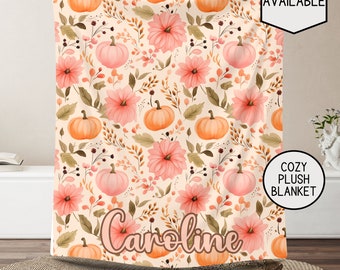May include: A cozy plush blanket with a fall-themed design featuring pink flowers, green leaves, and orange pumpkins. The blanket has the name "Caroline" written in a cursive font on the bottom.
