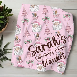 May include: Pink Christmas movie blanket with a Santa Claus, gingerbread man, and Christmas tree pattern. The blanket has the name "Sarah's" written on it.