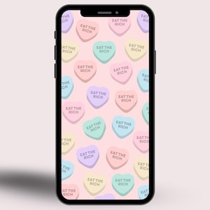 Puede incluir: Un smartphone muestra un fondo rosa con caramelos en forma de corazón de colores pastel repetidos. Cada caramelo tiene la frase "EAT THE RICH" impresa en blanco. El teléfono es negro y tiene una sombra.