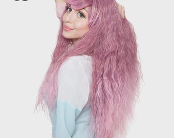 May include: A long, wavy, pastel pink wig with side-swept bangs. The hair features a textured, crimped appearance and falls past the shoulders. The top left corner displays the black text logo RockStar WIGS in a stylized, cursive font.