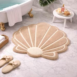 May include: A beige seashell-shaped bath mat with cream-colored accents. The mat is on a marble tile floor, near a white bathtub and a pair of beige slippers. A small white table holds bath products.